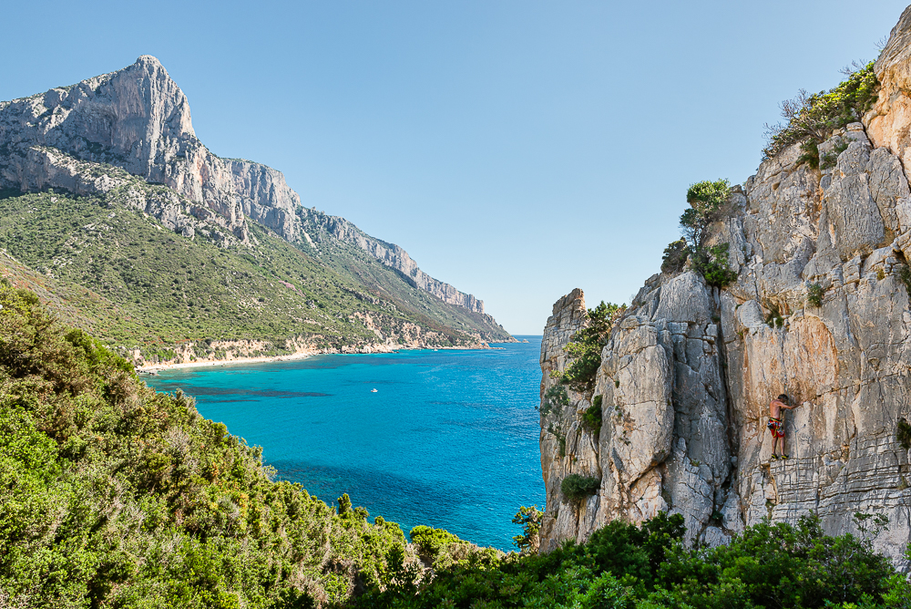 Ein Kletterer an den Felsen bei Pedra Longa mit dem Ausblick auf die Steilküste mit den Felswänden der Punta Giradili über dem türkisfarbenen Mittelmeer an der Ostküste von Sardinien, Ogliastra, Italien