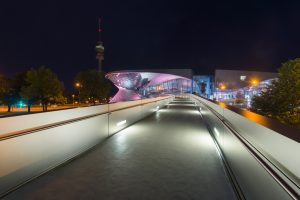 Beleuchtete Fassade der BMW Welt mit der Fußgängerbrücke vor dem Olympiaturm bei Nacht, München, Bayern, Deutschland