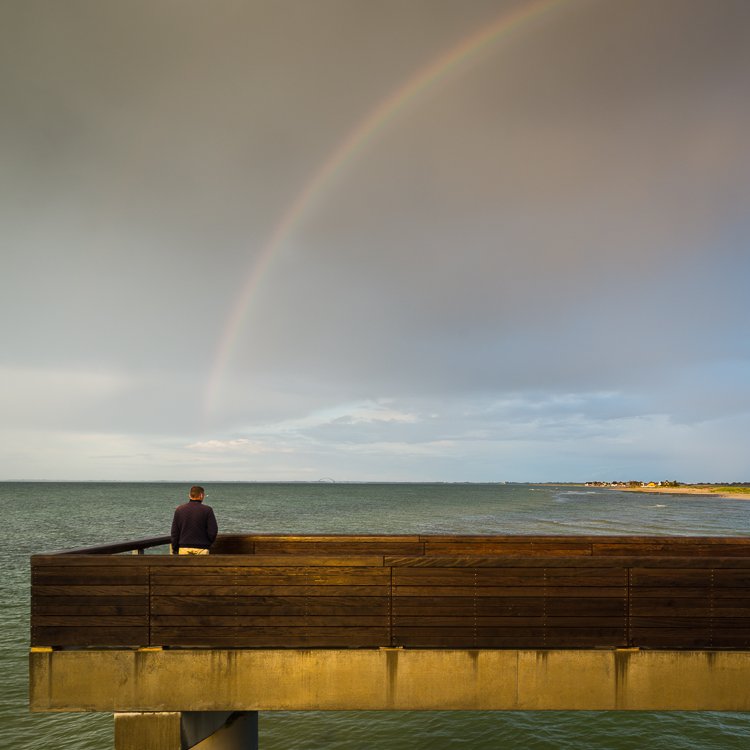 Mann auf der Seebrücke von Heiligenhafen beobachtet den Regenbogen über der Küste und der Halbinsel Graswarder