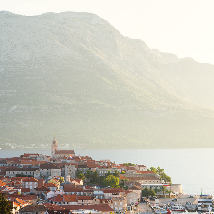 Die Altstadt von Korčula vor dem Gebirgsmasssiv des Sv. Ilija auf der Halbinsel Pelješac im Gegenlicht der Morgensonne, Insel Korčula, Süddalmatien, Kroatien