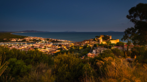 Blick über die Bastion, die Stadt und den Hafen von Castiglione della Pescaia auf eine weit geschwungene Bucht der Küste der Maremma in der Dämmerung, Toskana, Italien