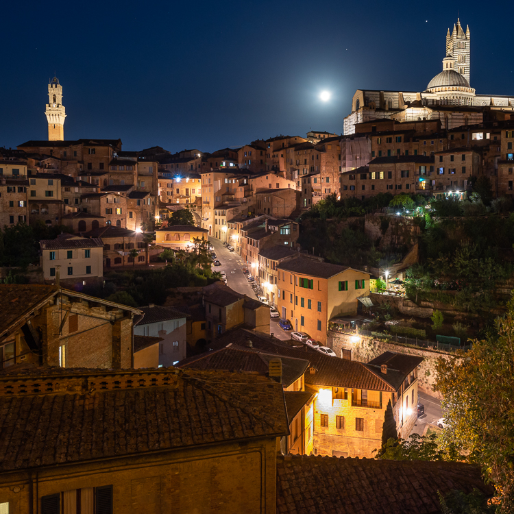 Vollmond über einer beleuchteten Gasse vor dem Panorama der mittelalterlichen Altstadt von Siena, Toskana, Italien