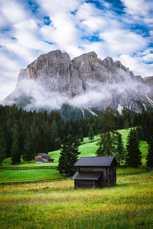 Heuhütten auf einer blühenden Almwiese vor dem Zwischenkofeltal und den Felswänden des Somamunt der Puez-Geisler-Gruppe, Campilltal Dolomiten, Südtirol, Italien
