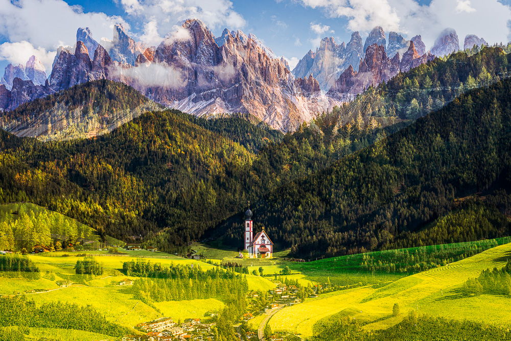 Herbstliche Landschaft der Dolomiten mit der Ranui-Kapelle in Villnöß vor Bergwäldern und den Felswänden der Geislerspitzen im Sonnenschein, Puez-Geisler-Gruppe, Südtirol, Italien