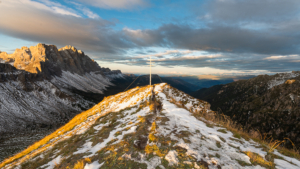 Gipfelkreuz des Zendleser Kofels mit den Geislerspitzen und der Villnößer Geisler Gruppe oberhalb des Villnößtals in den Dolomiten leuchtet im Sonnenschein bei Sonnenaufgang im Herbst, Südtirol, Italien