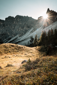 Die Strahlen der Morgensonne zwischen den Felsgipfeln der Puezspitzen bringen die Wiesen des Zwischenkofeltals unterhalb der Alm Antersasc zum Leuchten, Dolomiten Puez-Geisler-Gruppe, Südtirol, Italien