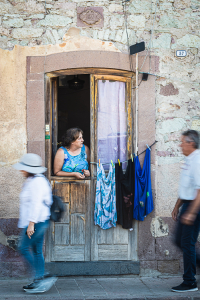 Sardinische Straßenszene - eine Frau schaut aus der Haustür nach rechts auf die Straße, während zwei Fußgänger auf dem Gehweg vorbeibergehen