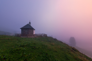 Das rötliche Licht des Sonnenuntergangs färbt die dicken Nebelschaden an der Kapelle der Ackernalm, Tirol, Österreich