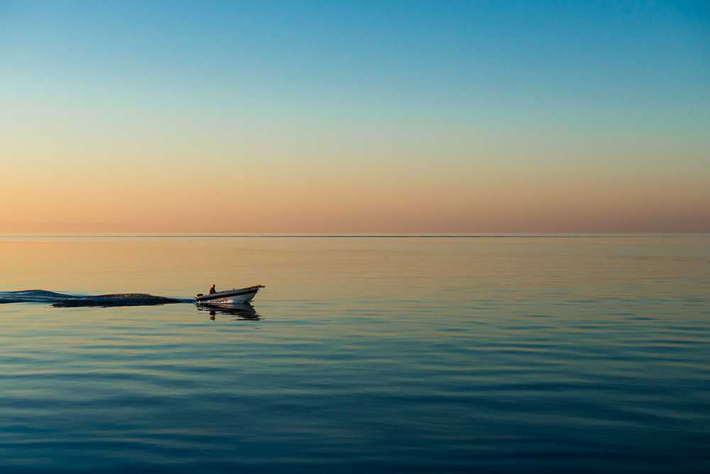 Ein Fischer fährt mit seinem Motorboot durch das stille Meer an der Küste von Jütland vor dem bunten Himmel der Morgendämmerung, Djursland, Dänemark