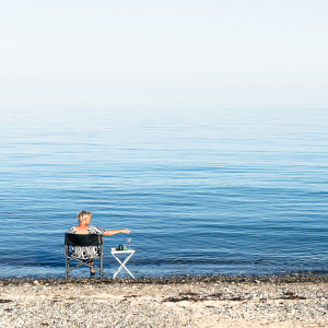 Eine Frau sitzt auf einem Campingstuhl am Ufer des Kattegatt in der Sonne und trinkt Wein, Djursland, Jütland, Dänemark