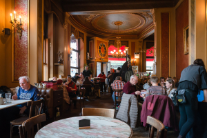 Der Gastraum im Wiener Kaffehaus Savoy mit verzierten Leuchtern und einer Decke mit runden und vergoldeten Stuckelementen