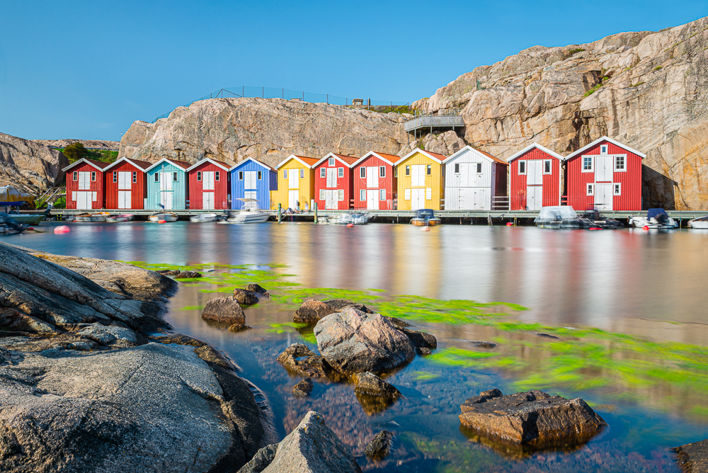 Smögenbryggan: Bootsschuppen und Speicherhäuser mit bunten Holzfassaden zwischen Granitfelsen an der Hafenpromenade von Smögen in den Schären der schwedischen Westküste bei Sonnenschein