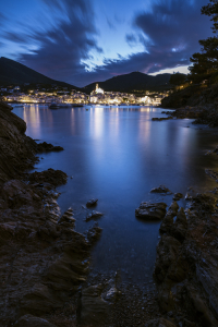 Blick von einer felsigen Bucht auf die beleuchtete Altstadt von Cadaqués in der Abenddämmerung, Katalonien, Spanien