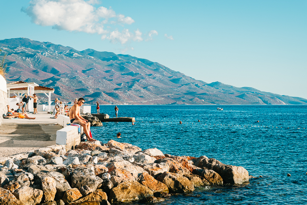 Badegäste an einer Strandbar in Senj an der kroatischen Adriaküste mit dem Velebit-Gebirge in der Abendsonne Badegäste am Ufer an einer Strandbar in Senj an der kroatischen Adriaküste mit dem Velebit-Gebirge in der Abendsonne
