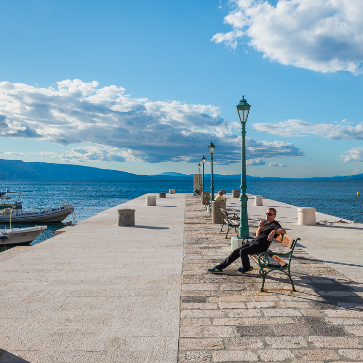 Menschen sitzen in der Sonne auf Bänken im Hafen von Senj mit Blick auf die Insel Krk in der Kvarner Bucht von Kroatien Menschen sitzen in der Sonne auf Bänken im Hafen von Senj mit Blick auf die Insel Krk in der Kvarner Bucht von Kroatien